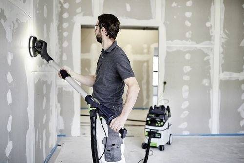 Een persoon gebruikt een schuurmachine op gipsplaat in een kamer die in aanbouw is, met een stofzuiger aangesloten voor stofafzuiging.
