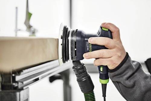 Een persoon gebruikt een elektrische schuurmachine op een houten oppervlak, waarbij hij zich richt op het gladmaken van de rand. De schuurmachine is zwart en groen, met een slang aangesloten.