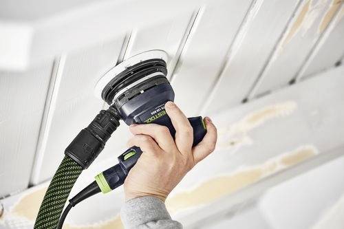 Een persoon gebruikt een elektrische schuurmachine om een houten plafond glad te schuren, waardoor plekken van lichter hout zichtbaar worden onder de oude verflaag.