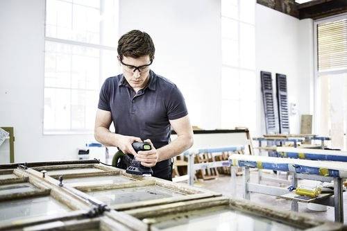 Een persoon die veiligheidsbril draagt, werkt aan houten raamkozijnen in een werkplaats, waarbij hij gebruikmaakt van een elektrisch gereedschap. Heldere ramen verlichten de ruimte.
