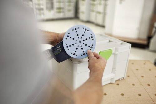 Een persoon monteert een elektrische schuurmachine, waarbij hij een blauwe schuurschijf met gaten op het gereedschap plaatst, in een werkplaatsomgeving met gereedschappen op de achtergrond.