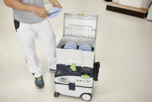 Een persoon die witte broek draagt, bedient een professionele schoonmaakmachine en plaatst schijven erin. De vloer en verschillende mogelijke apparaten zijn zichtbaar.