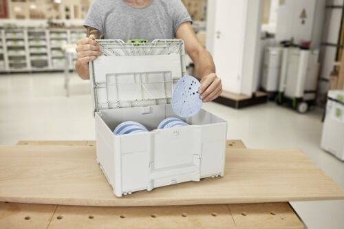 Een persoon plaatst een rond, geperforeerd voorwerp in een witte opslagdoos die soortgelijke items bevat, in een heldere, georganiseerde werkruimte.
