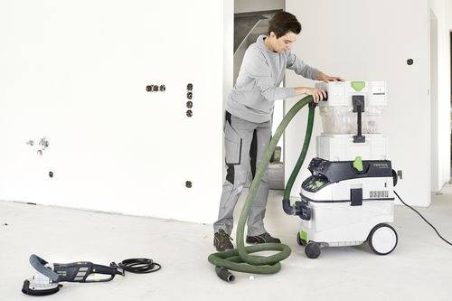 Een persoon in werkkleding stelt een slang bij op een grote industriële stofzuiger in een onbemeubileerde ruimte. Een afzonderlijke elektrische gereedschap ligt op de vloer.