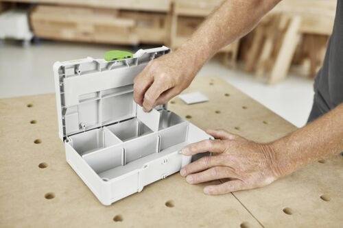 Een persoon die een witte opslagdoos met scheidingswanden organiseert op een geperforeerde houten tafel, vermoedelijk in een werkplaatsomgeving.