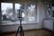 Een op een statief gemonteerde camera met een helder licht staat in een lege ruimte met grote ramen, wat wijst op een focus op fotografie of filmen.