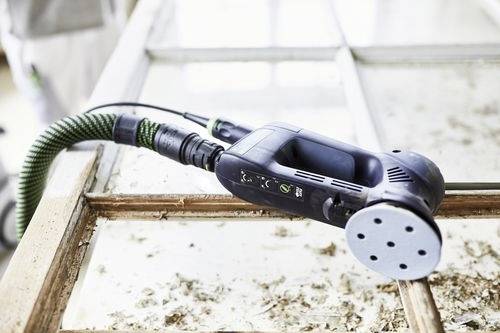 Een elektrische schuurmachine met een groene slang rust op een raamkozijn, waarbij oude verf en vernis worden verwijderd met houtkrullen verspreid rond de machine.