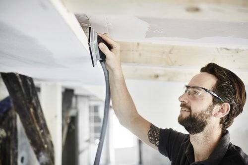 Een persoon die veiligheidsbril draagt, schuurt een plafond met een handgereedschap in een werkplaats, met de focus op het gladmaken van een houten balk.