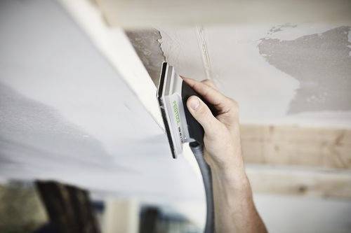 Een persoon gebruikt een handschuurmachine aan het plafond, waarbij hij het pleisterwerk glad maakt. Licht valt binnen door een raam en verlicht de werkruimte.