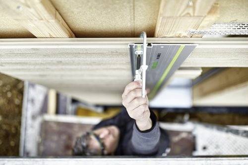 Een persoon installeert een houten paneel met behulp van een railsysteem, gezien vanuit bovenaanzicht. De focus is op het paneel en de rail, waarbij precisiewerk wordt getoond.