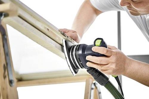 Een persoon gebruikt een elektrische schuurmachine op een houten oppervlak, met grote concentratie. De omgeving doet denken aan houtbewerking of woningverbetering.