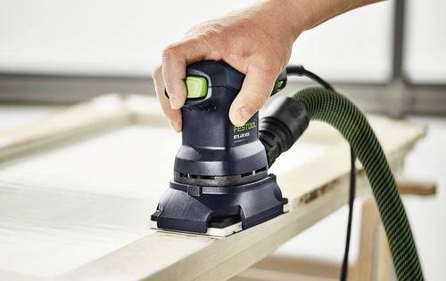 Een persoon gebruikt een kleine elektrische schuurmachine op een houten oppervlak, met een groene slang aangesloten voor stofafzuiging.