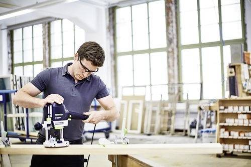 Een man die een elektrisch gereedschap gebruikt op een houten plank in een werkplaats. Hij draagt veiligheidsbril, en de omgeving toont grote ramen en schappen.