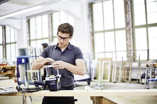 Een man in een werkplaats stelt een gereedschap bij op een werkbank, omringd door grote ramen en houten frames, waarbij een houtbewerkingsomgeving wordt uitgebeeld.