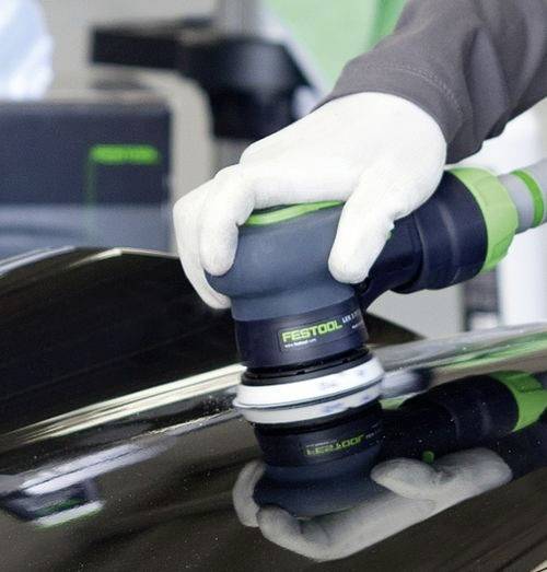 'Gehandschoende handen die een Festool excentrische schuurmachine gebruiken op een glanzend zwart oppervlak, waarbij een gladde afwerking wordt gecreëerd in een werkplaatsomgeving.'