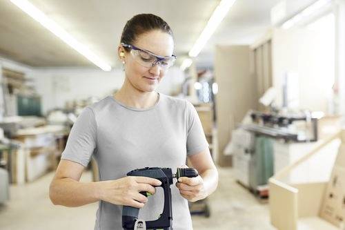 Een persoon met veiligheidsbril en oordopjes bedient een elektrisch gereedschap in een werkplaatsomgeving, wat een nadruk op veiligheid op de werkplek aangeeft.