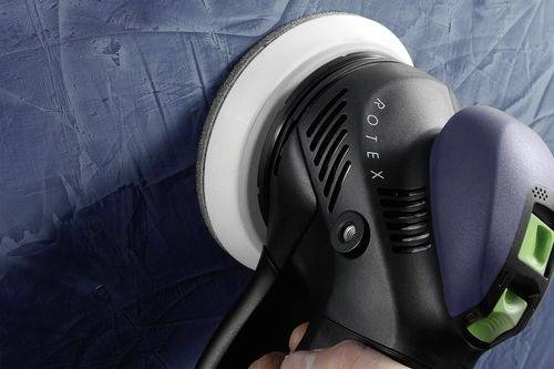 Een hand houdt een elektrische schuurmachine van het merk 'Rotex' vast, waarbij een gestructureerd blauw oppervlak wordt gladgeschuurd, wat wijst op een stap in een doe-het-zelf- of reparatieproject.