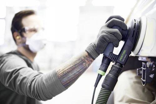 Een persoon met veiligheidsbril en masker gebruikt een elektrische schuurmachine op het oppervlak van een voertuig, waarbij de nadruk wordt gelegd op veiligheid en precisie in automotive werkzaamheden.