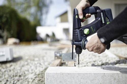 Een persoon die een boormachine gebruikt op een betonnen blok in de buitenlucht, met een onscherpe achtergrond van constructielocatie-elementen.