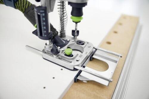 Een boormachine is bevestigd aan een werkbank, weergegeven in gebruik met een groen en zwart handvat. Hij is gepositioneerd om nauwkeurig boren in hout te geleiden.