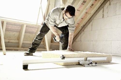 Een man gebruikt een elektrisch gereedschap om een stuk hout te zagen in een zolder in aanbouw, waarbij een klus voor woningverbetering wordt getoond.
