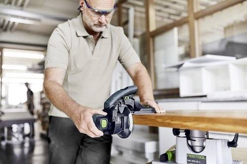 Een persoon met veiligheidsbril gebruikt een elektrisch gereedschap om een houten plank te schuren in een werkplaatsomgeving.