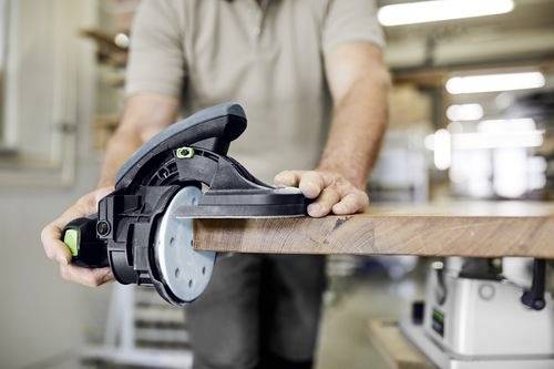 Een persoon gebruikt een vlakschuurmachine aan de rand van een houten plank, met de focus op precisie en het gladmaken van het oppervlak in een werkplaatsomgeving.