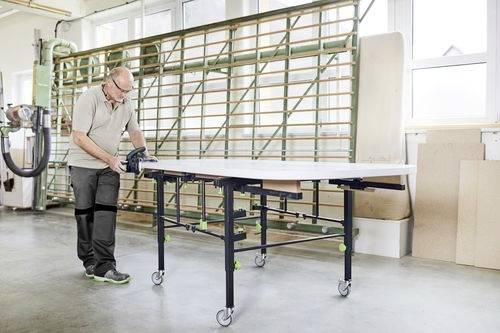 Een persoon meet een groot bord op een verrijdbare tafel in een helder verlichte werkplaats, met apparatuur en opslag zichtbaar op de achtergrond.