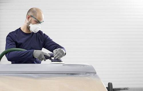 Een persoon in beschermende uitrusting gebruikt een schuurgereedschap op het oppervlak van een voertuig, met de focus op een specifiek gedeelte in een werkplaatsomgeving.