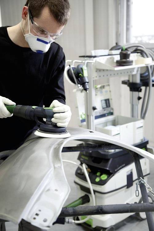 'Persoon in beschermende uitrusting die een schuurmachine gebruikt op een autoonderdeel in een industriële omgeving. Apparatuur en gereedschappen zichtbaar op de achtergrond.'