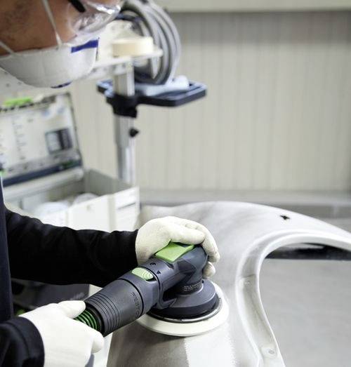 Persoon met beschermende uitrusting gebruikt een elektrisch gereedschap om een auto-bumper te polijsten in een werkplaatsomgeving.