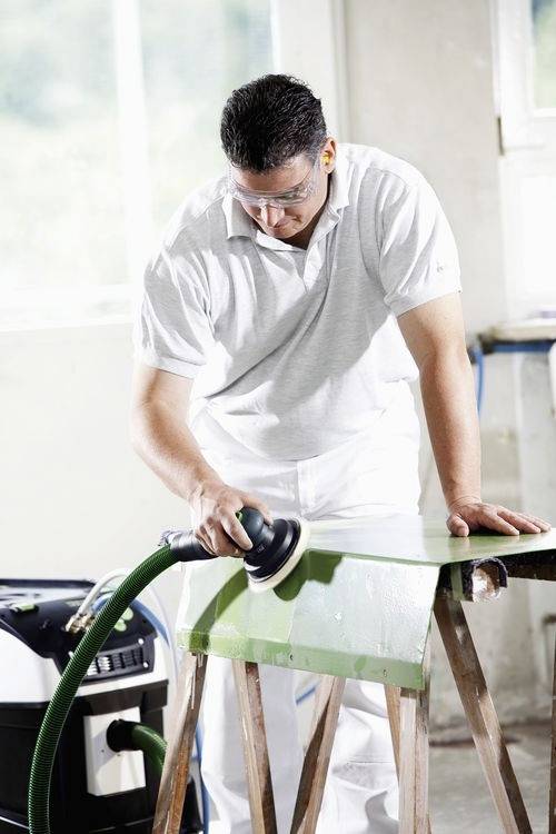 Een persoon die veiligheidsbril en werkkleding draagt, schuurt een groene houten plank met een elektrische schuurmachine in een werkplaatsomgeving.