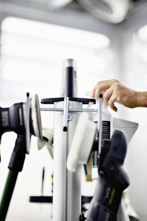 Een persoon past gereedschappen aan op een standaard in een werkplaats, met de focus op organisatie en bereikbaarheid van gereedschappen. Verschillende uitrustingsstukken zijn zichtbaar op de achtergrond.