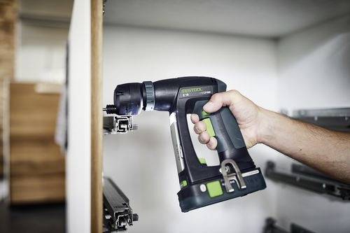 Een persoon gebruikt een draadloze boormachine om hang- of montagemateriaal te installeren of bij te stellen op een wit kastje of een stellingkast. De scène speelt zich binnenshuis af.