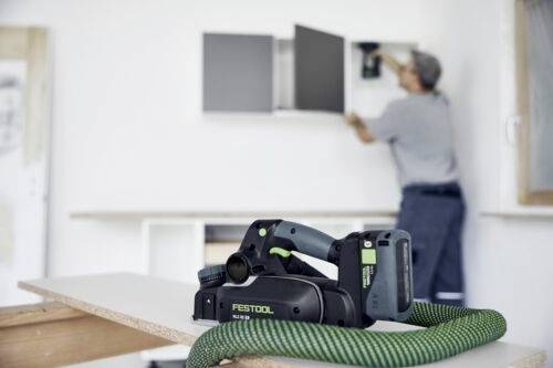 Een draadloos elektrisch gereedschap met een slang ligt op een tafel; een persoon op de achtergrond bevestigt planken aan een witte muur.