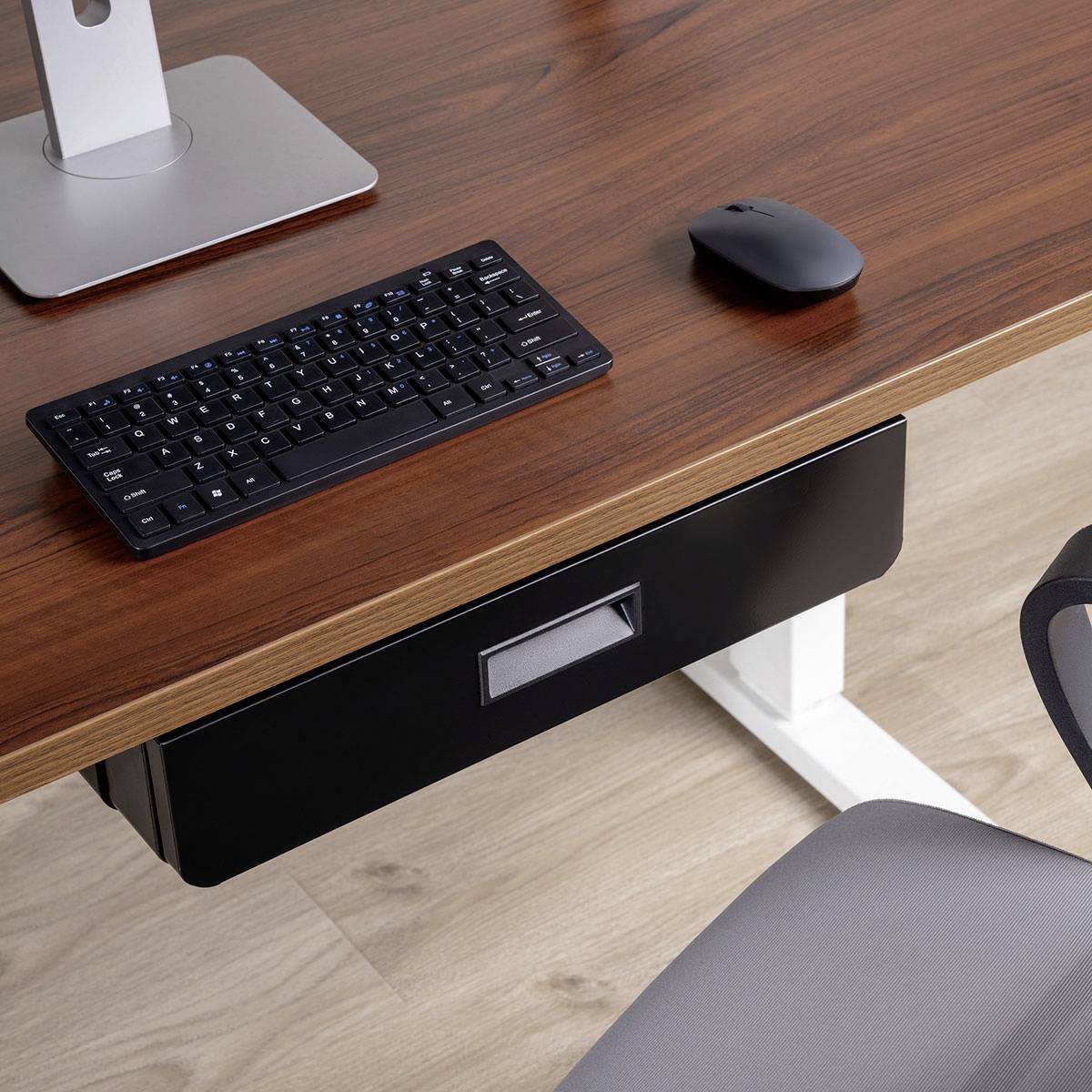 Een houten bureau met een zwart toetsenbord en muis. Een zilveren standaard staat links. Een grijze bureaustoel is gedeeltelijk zichtbaar onderaan.