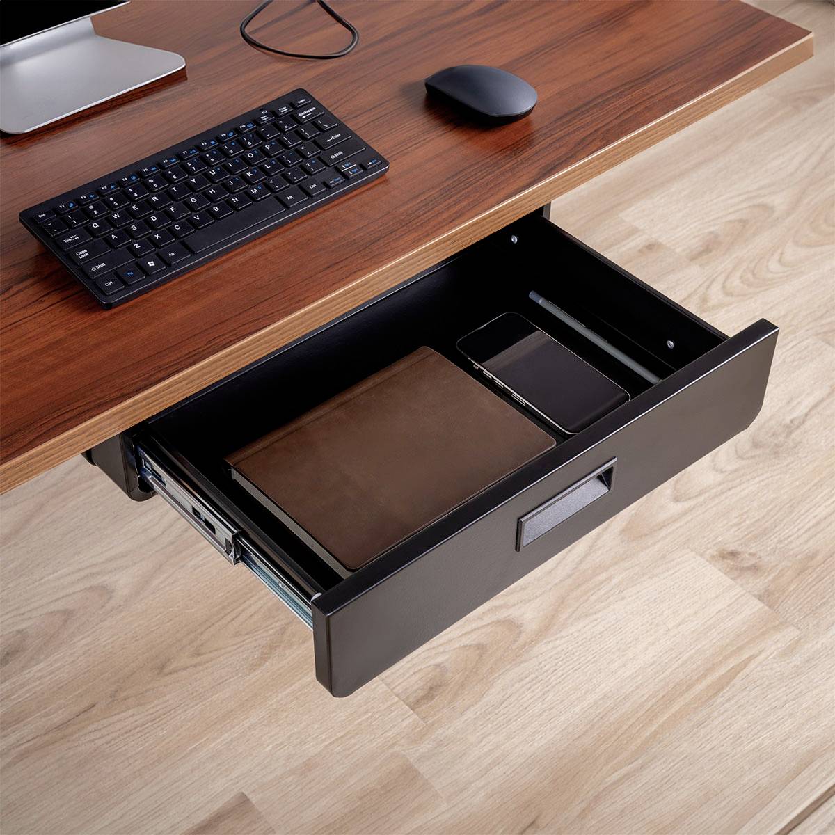 Een open bureaulade onthult een gesloten laptop, een bruine notebook en een tablet aan de binnenkant. Een toetsenbord en muis bevinden zich op het bureau erboven.