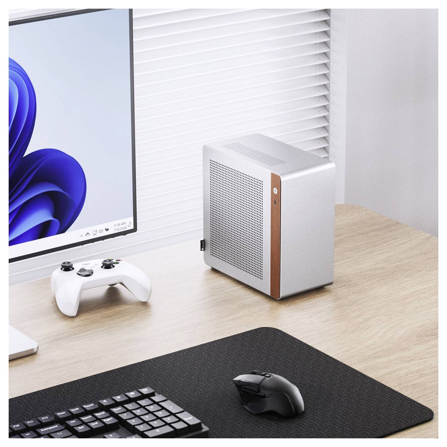 Een minimalistische werkruimte met een compacte, zilveren desktopcomputer naast een monitor, met een witte gamingcontroller en toetsenbord op een houten bureau.