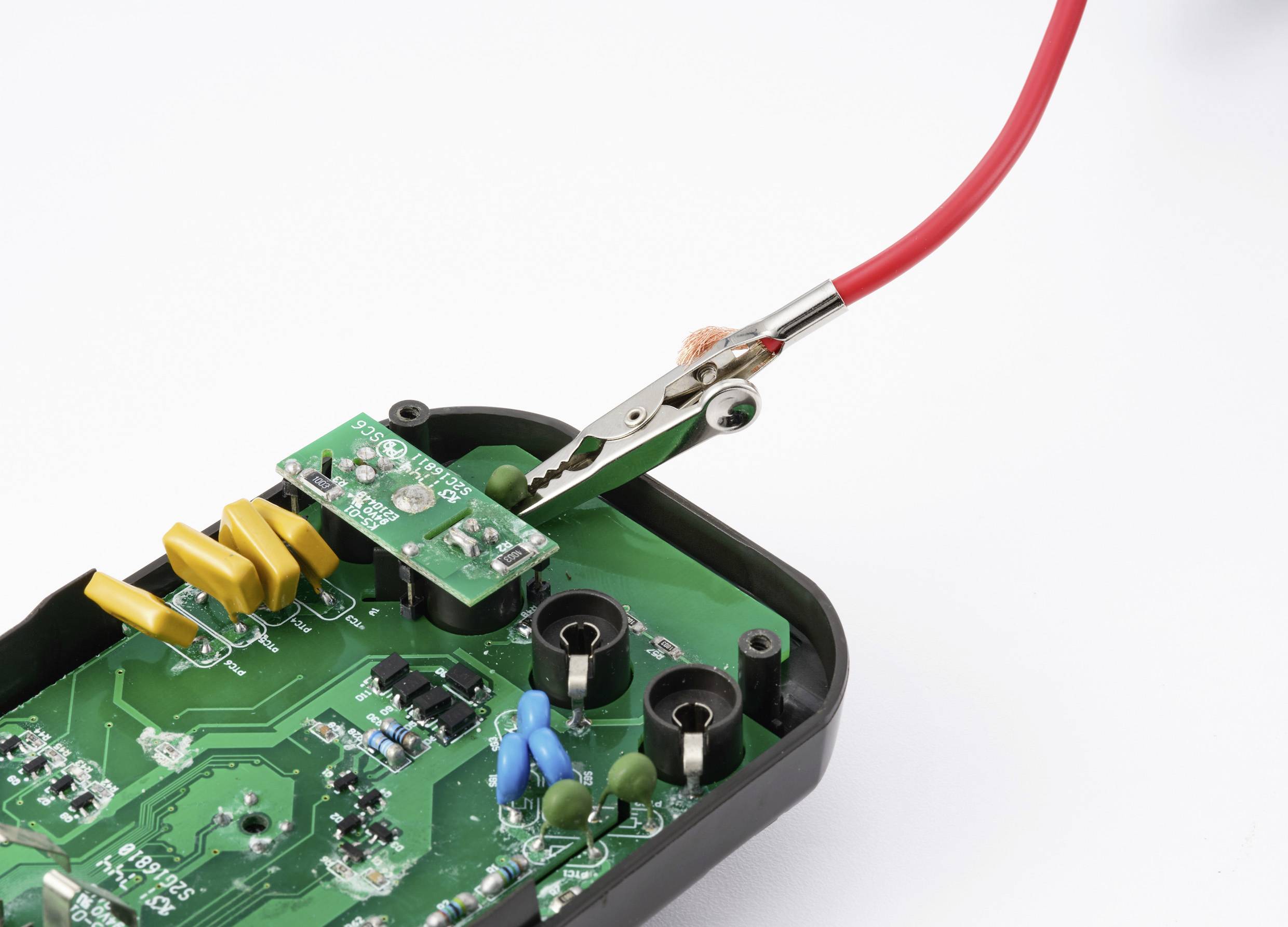 Een printplaat met een testclip en een rode draad aangesloten, waarop kleine componenten en groene borddetails zichtbaar zijn, gebruikt voor elektronisch testen.