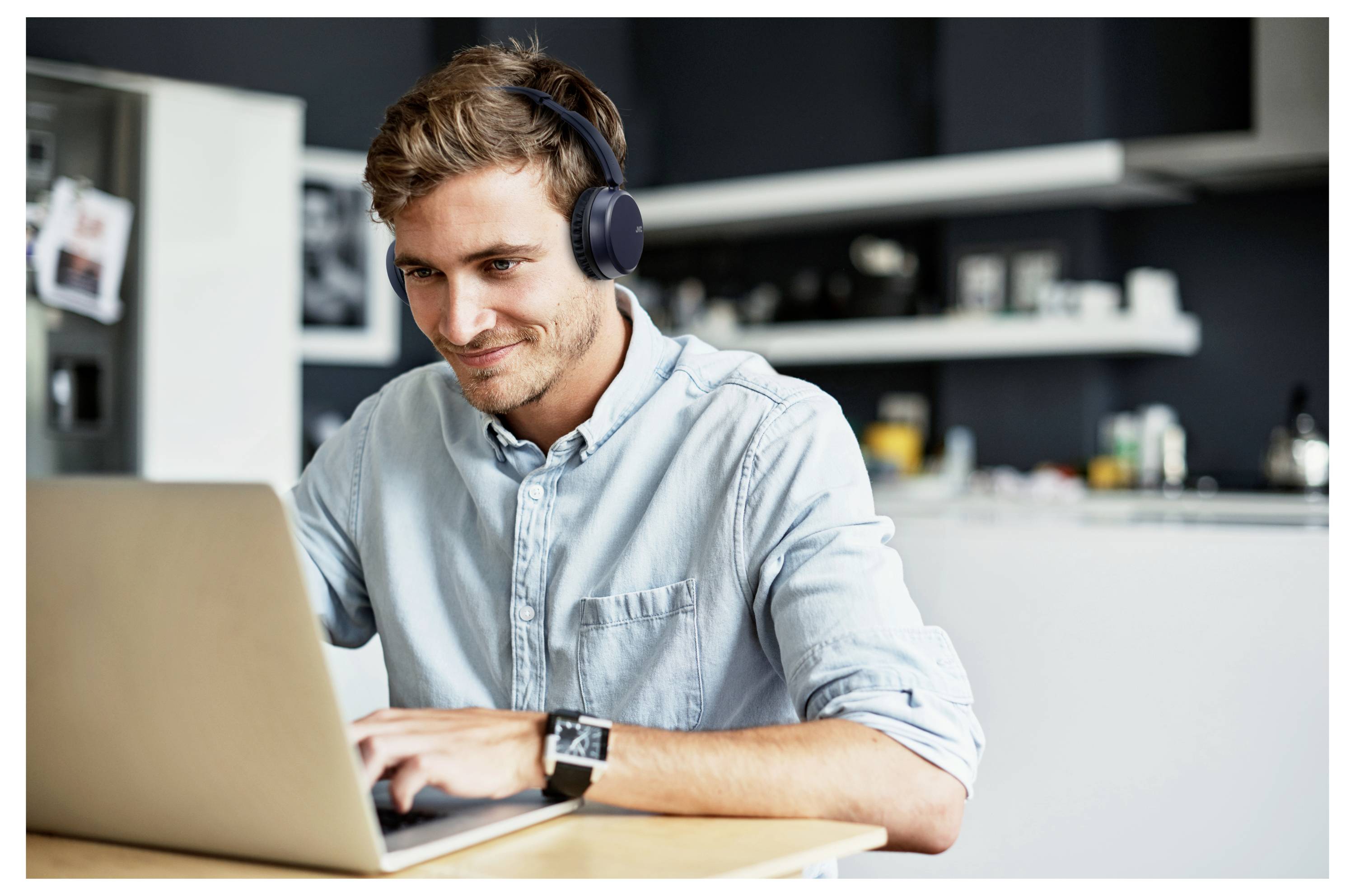 Een man in een casual overhemd zit aan een tafel, werkt op een laptop en draagt koptelefoons, in een moderne keukenomgeving.