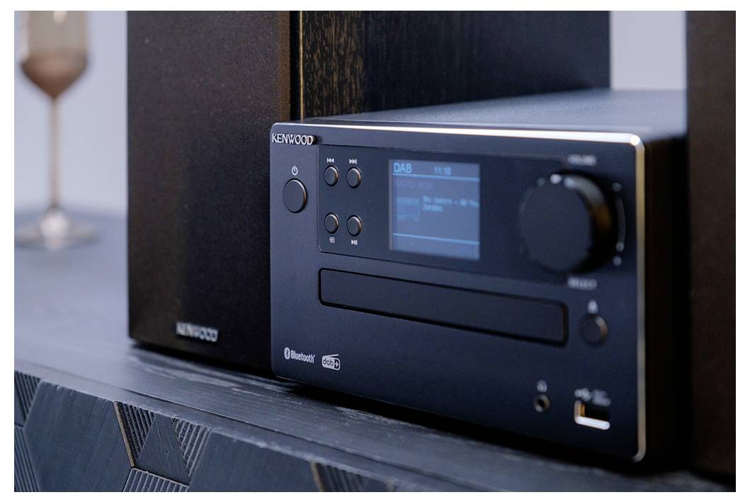 Een stereosysteem met een digitaal display waarop het radiostation wordt weergegeven, een volumeknop en een cd-sleuf, geplaatst op een moderne plank naast speakers.
