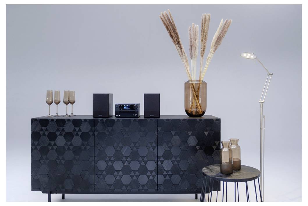 Donker geometrisch dressoir met wijnglazen en een stereo. Bruine vazen met pampasgras bovenop. Een staande lamp en bijzettafel ernaast.