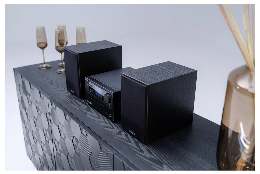 Een zwart stereosysteem met twee speakers op een gestructureerde kast, omringd door drie bruingetinte wijnglazen en een decoratieve vaas.