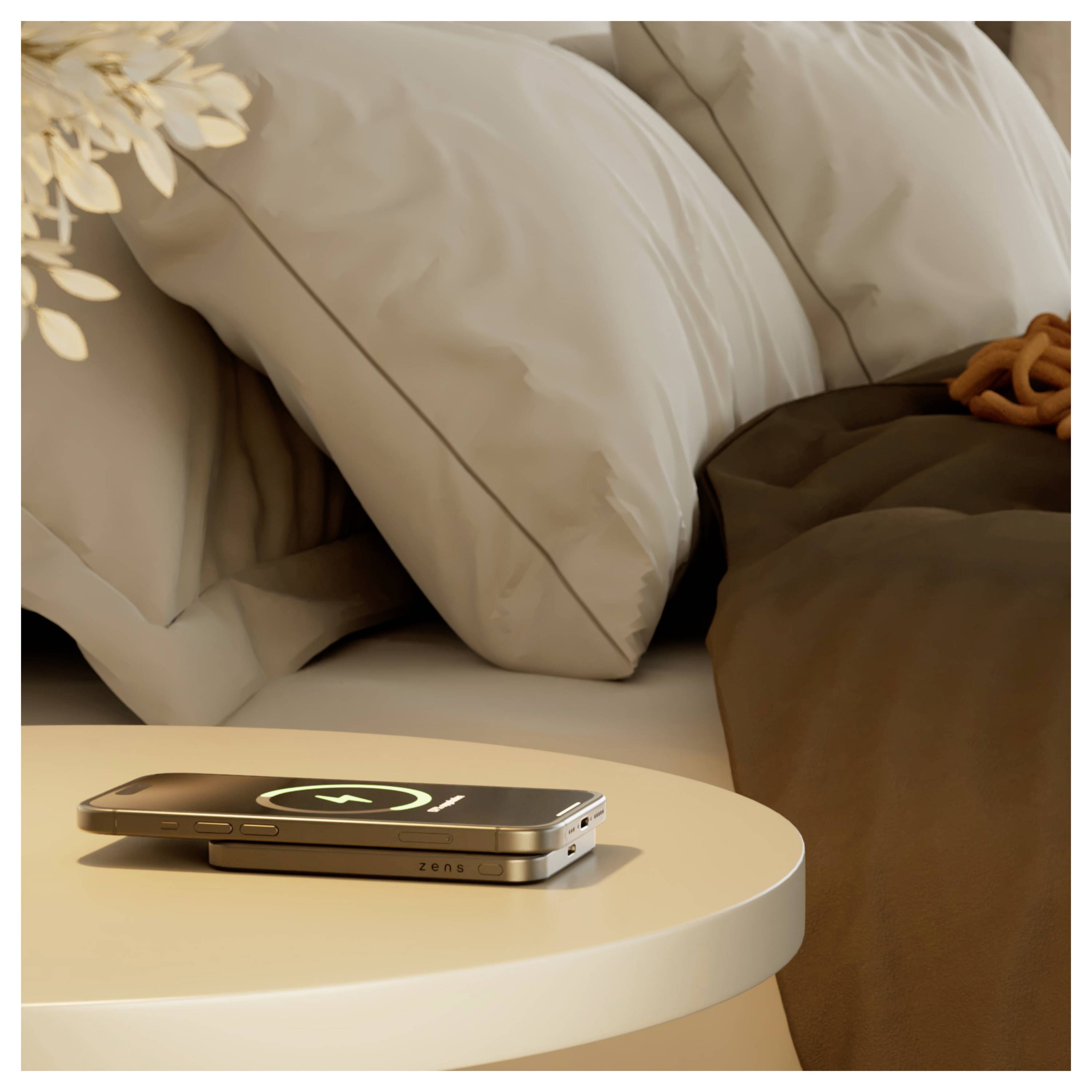 Een smartphone met een oplaadsymbool rust op een rond nachtkastje naast een netjes opgemaakt bed met witte kussens en een bruine deken.