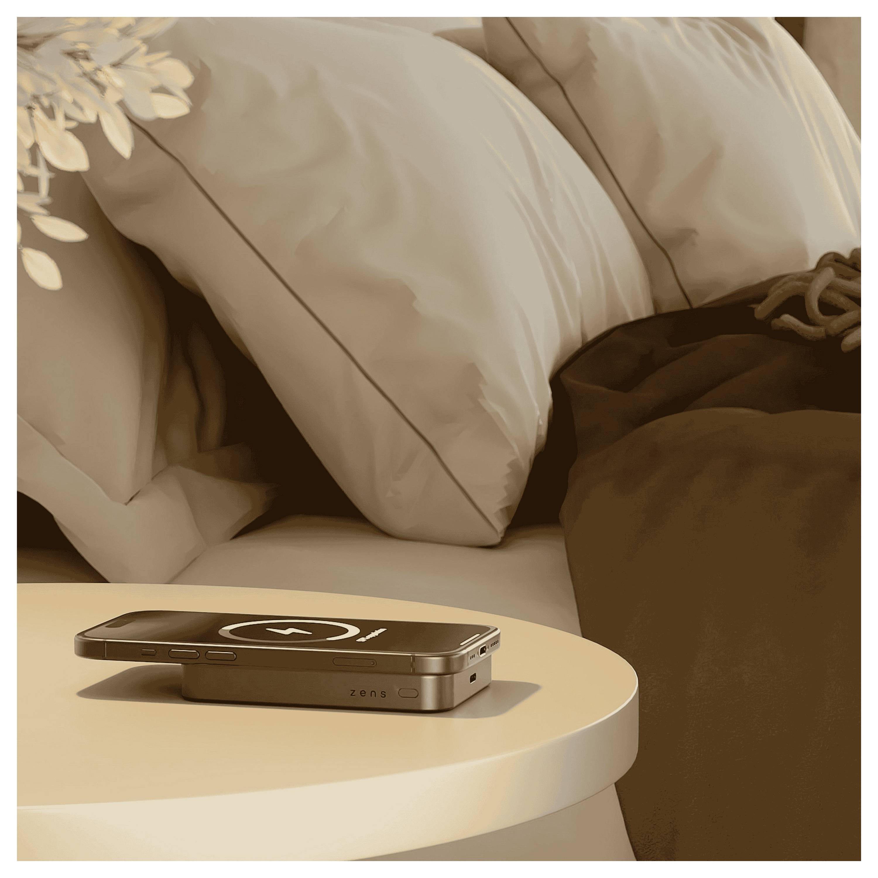 Een smartphone met een oplaadpictogram rust op een rond nachtkastje naast een bed met kussens en dekens, wat suggereert dat het apparaat klaar wordt gezet voor het opladen voordat men gaat slapen.