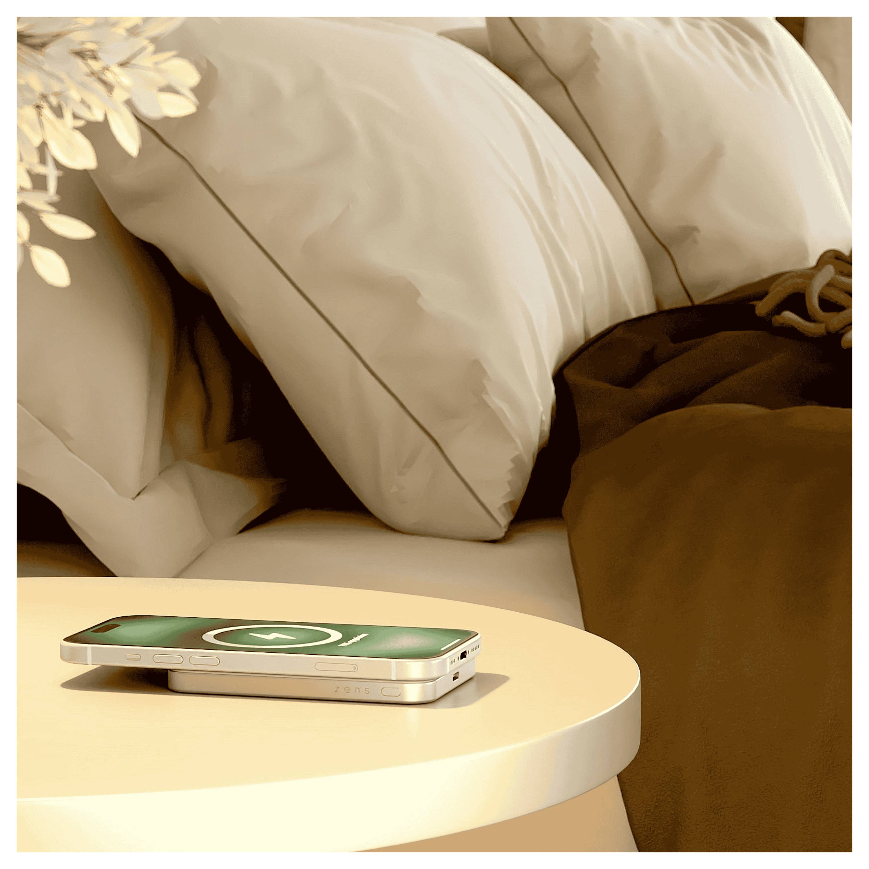 Een smartphone met een groen batterijlaadpictogram rust op een nachtkastje naast een netjes opgemaakt bed met witte kussens en bruine lakens.