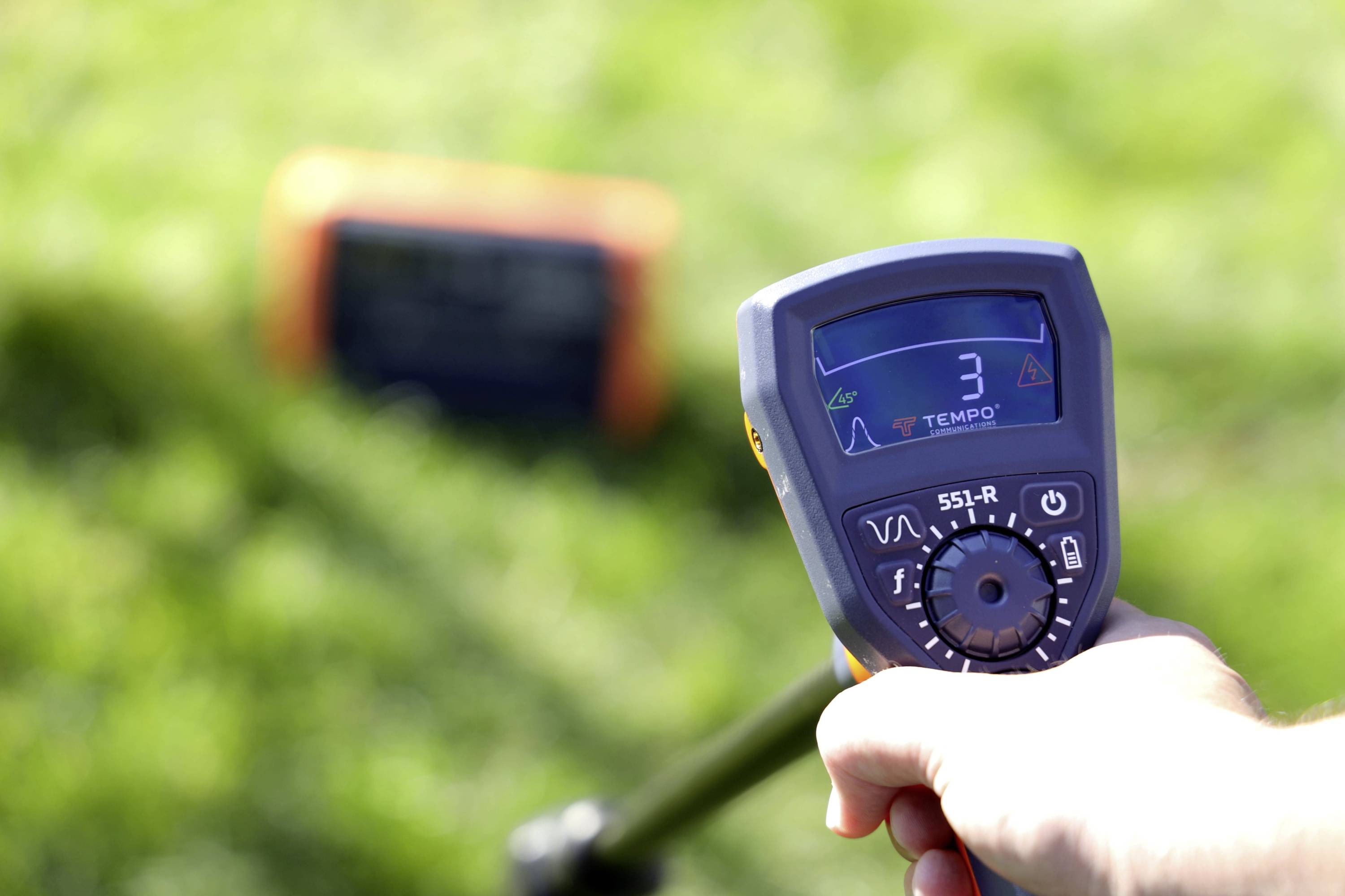 Een hand houdt een Tempo-apparaat vast met een scherm waarop het getal '3' wordt weergegeven, gericht op een oranje object op gras, wat wijst op meting of detectie.