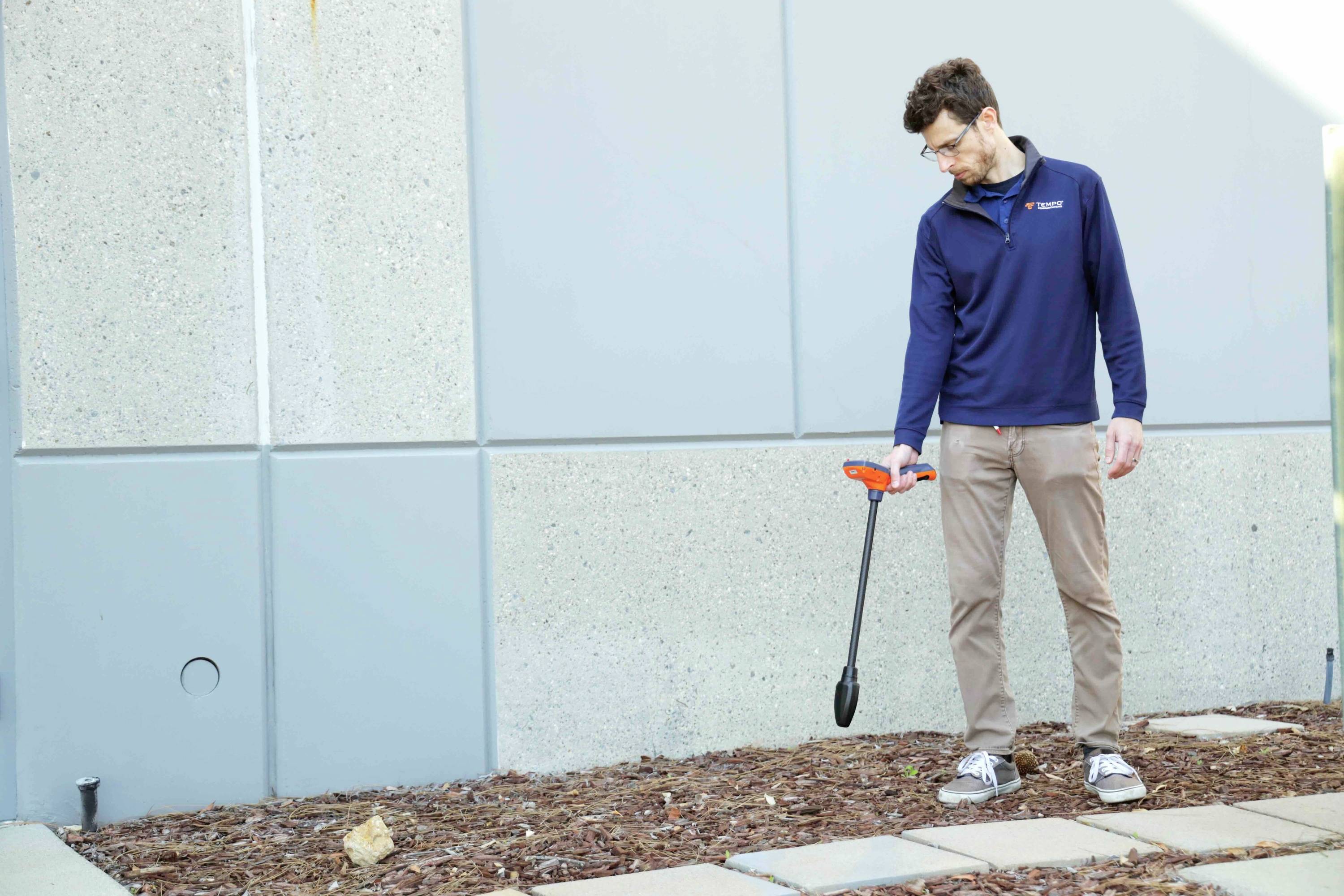 Een man in vrijetijdskleding gebruikt een metaaldetector om de met houtsnippers bedekte grond nabij de betonnen muur van een gebouw te doorzoeken.