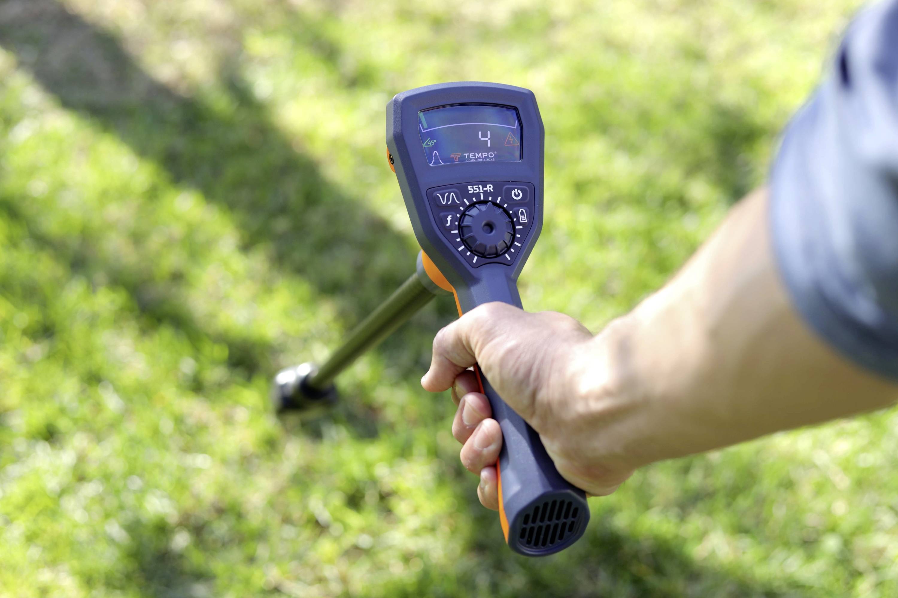 Een persoon houdt een metaaldetector boven graszodegrond en scant naar begraven voorwerpen. Het display toont '4' met verschillende instellingen zichtbaar.