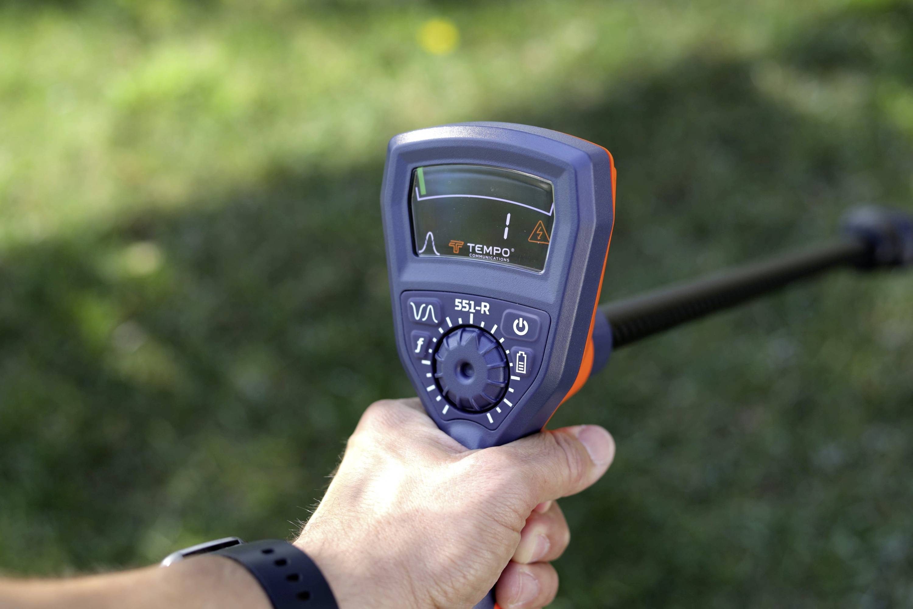 Een hand die een apparaat vasthoudt met een digitaal scherm en meerdere draaiknoppen, gebruikt voor veldmetingen of detectie, met gras op de achtergrond.